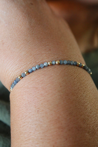 3mm sapphire gemstone gold bead bracelet on a wrist with a blurred green fabric t shirt background